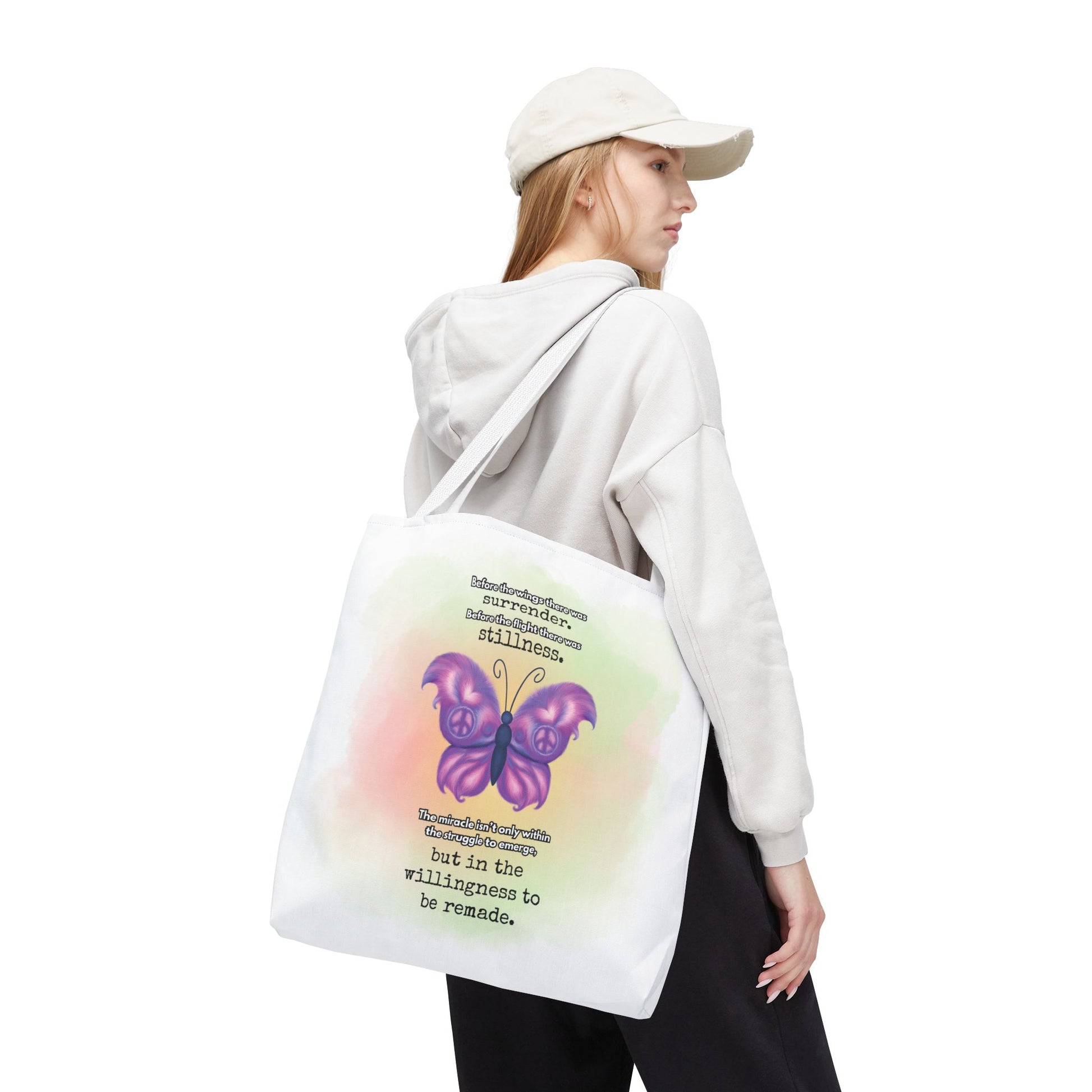 Person wearing a white hoodie and cap, holding a tote bag with a butterfly design and text.