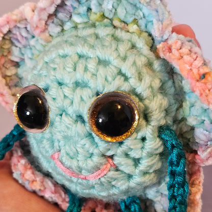 Close-up of Floyd Flower Boy’s mint crochet face with large gold-rimmed sparkly eyes, a small pink smile, and pastel flower petals.