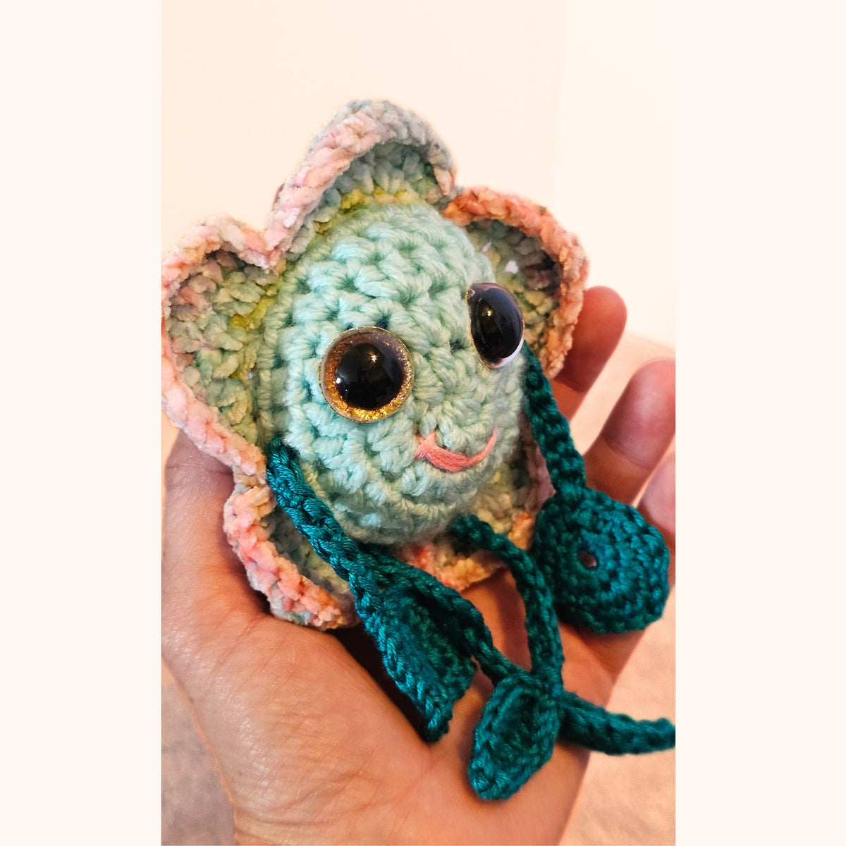 Handheld angled view of Floyd Flower Boy showing his pastel crochet flower petals, mint face, sparkly eyes, and dangling teal limbs.