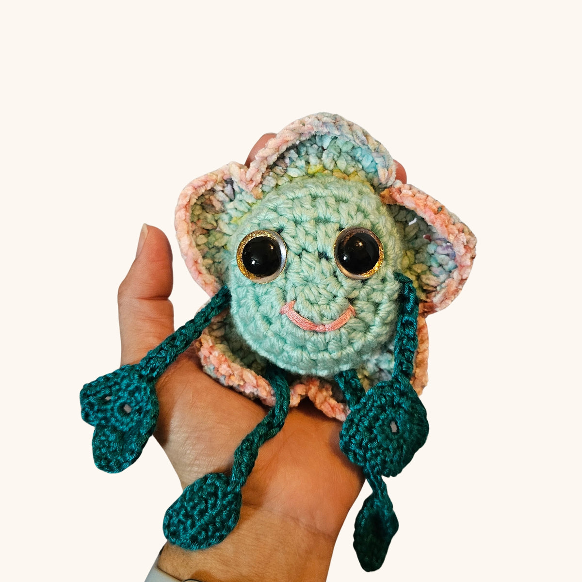 Floyd Flower Boy resting in a hand to show the scale of the handmade crochet keepsake, with pastel petals and dangling teal limbs.