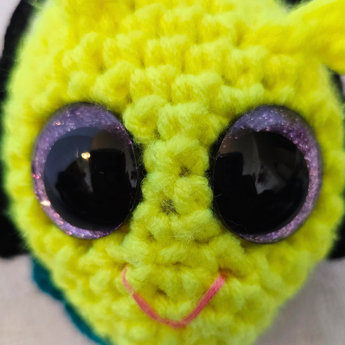 Close-up of Groovy Gus with a neon yellow-green crochet face, large purple-rimmed sparkly eyes, small antenna-like bumps, and pink stitched smile.