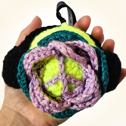 Handheld back view of Groovy Gus showing the lavender crochet peace sign layered over teal, yellow-green, and black yarn.