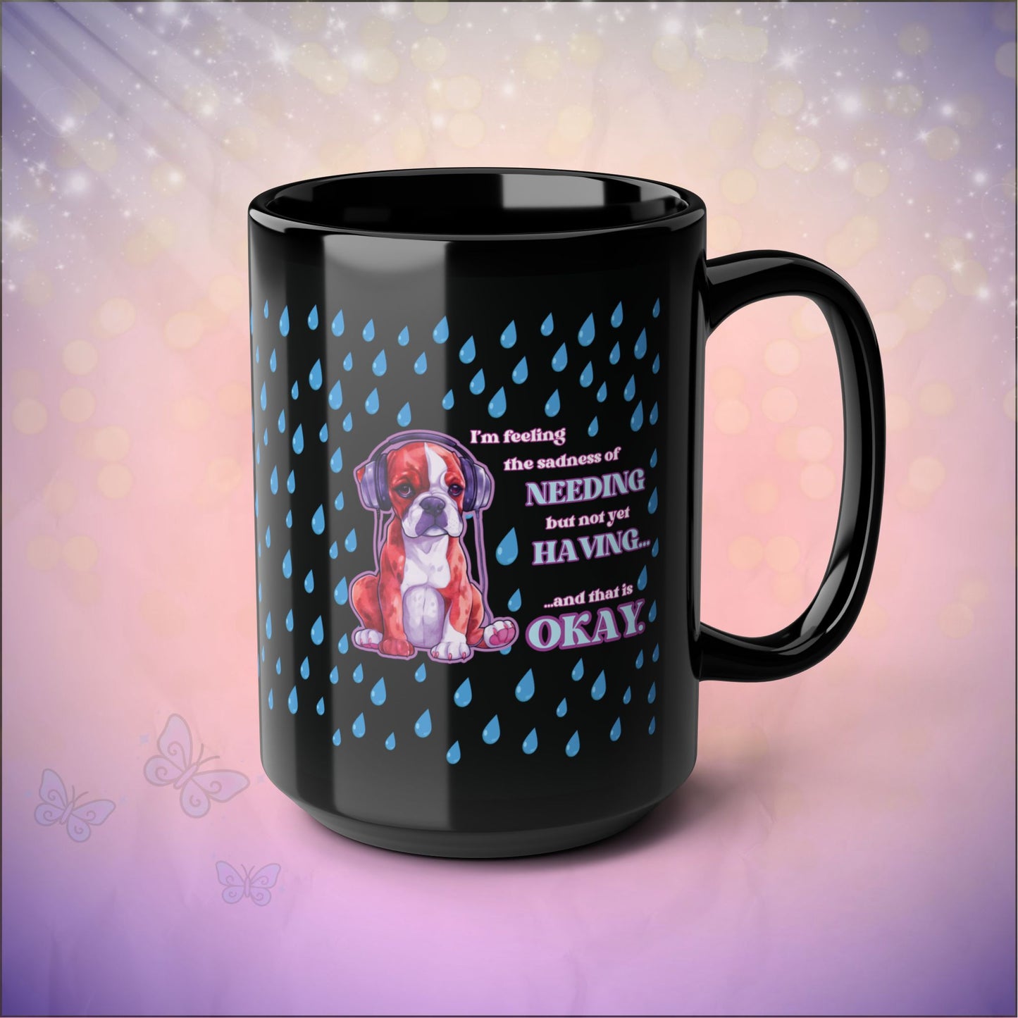 Black 15 oz ceramic mug with a glossy finish featuring a sad boxer puppy wearing headphones beneath blue raindrops. The quote reads, “I’m feeling the sadness of needing but not yet having… and that is okay.” The mug sits on a soft pink and purple gradient background with sparkles and butterfly accents.