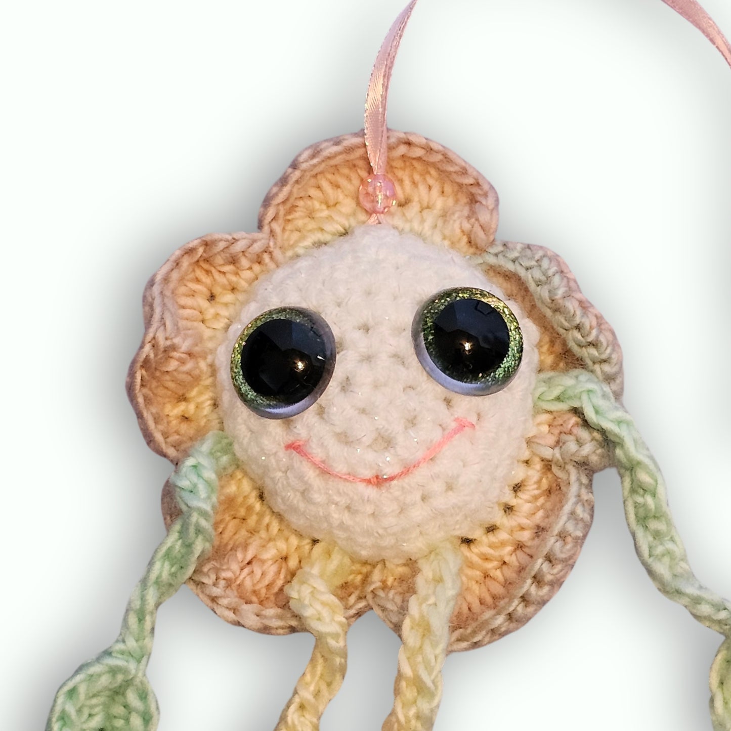 A handmade crocheted flower ornament with a smiling face, large shiny black eyes, and a pink hanging ribbon, shown against a plain white background.