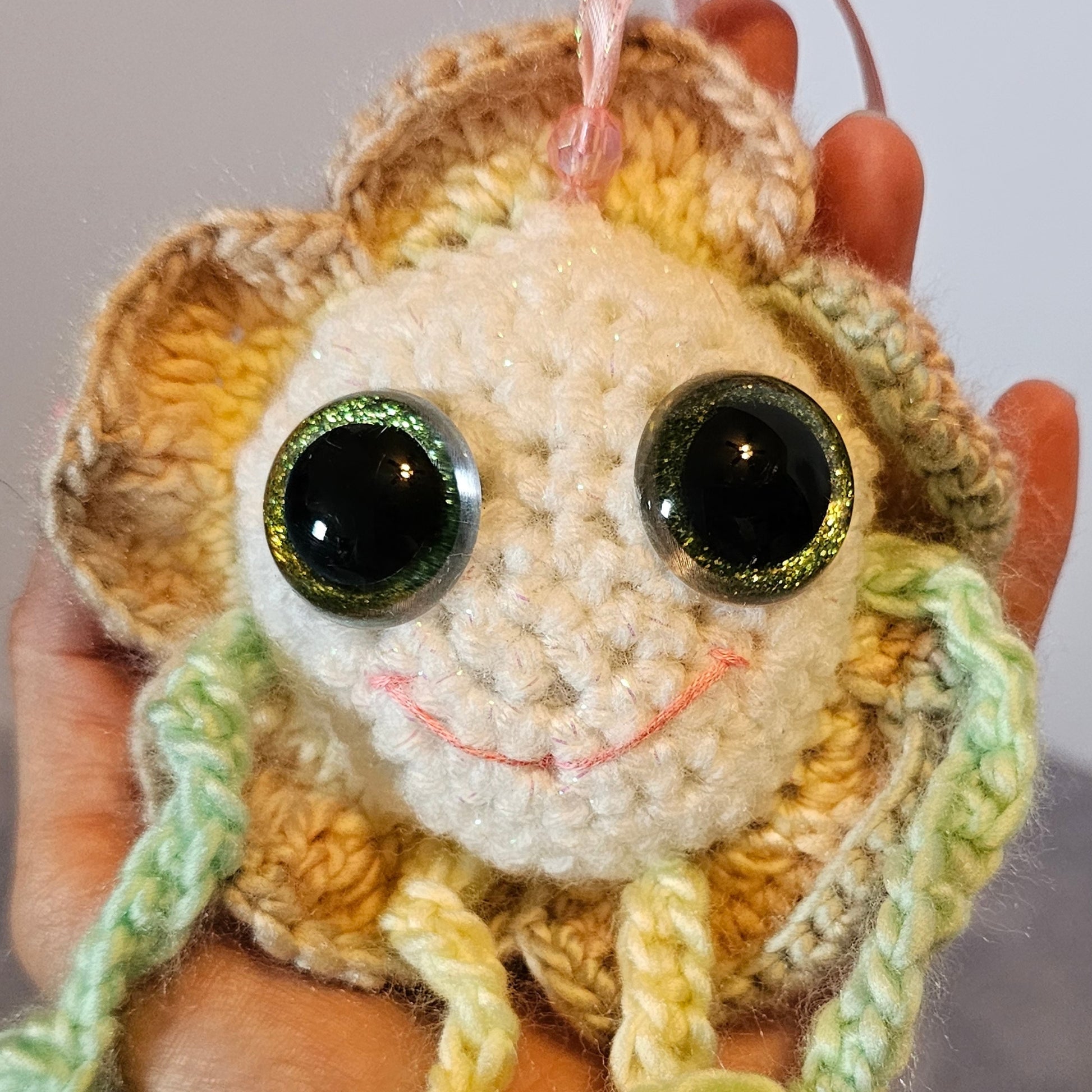 A close-up of a hand holding a crocheted jellyfish toy. The toy has a cream-colored face, a yellow and brown textured body, green and yellow tentacles, and large shiny black eyes.