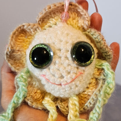A close-up of a hand holding a crocheted jellyfish toy. The toy has a cream-colored face, a yellow and brown textured body, green and yellow tentacles, and large shiny black eyes.
