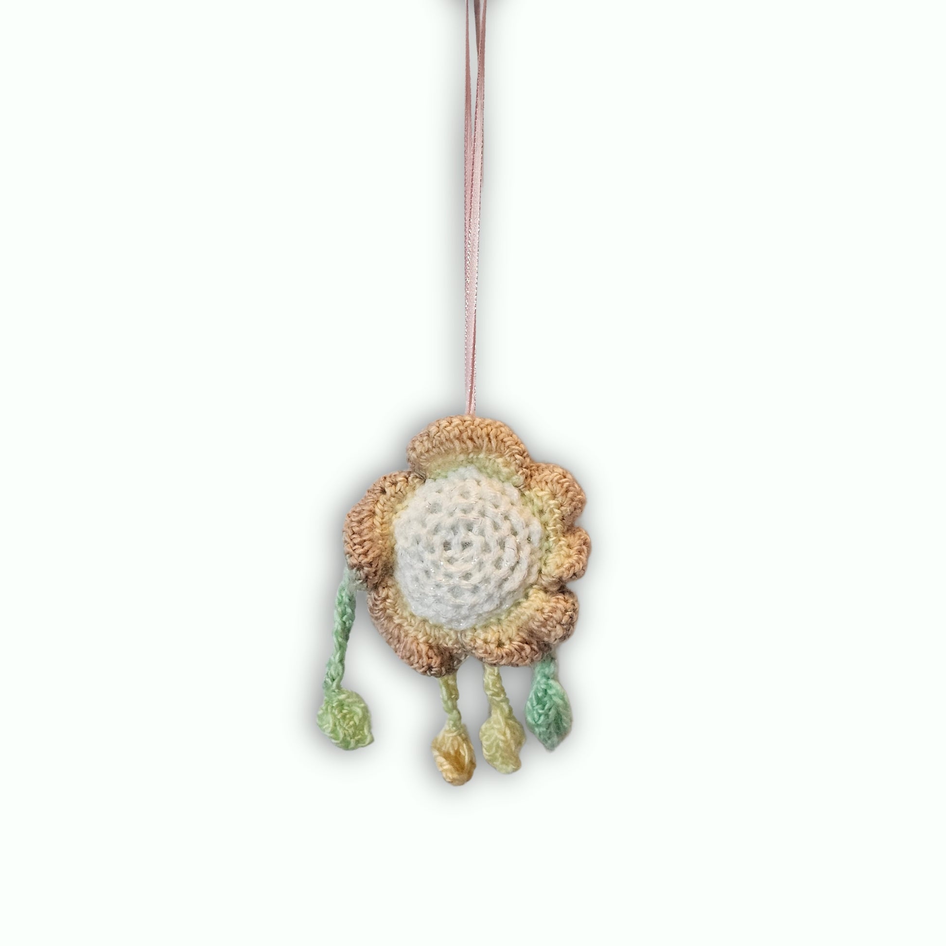 A handmade crocheted flower ornament with a beige and brown textured outer ring, a white center, and three hanging tassels in green, yellow, and beige, suspended by a thin brown string against a white background.