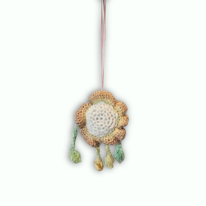 A handmade crocheted flower ornament with a beige and brown textured outer ring, a white center, and three hanging tassels in green, yellow, and beige, suspended by a thin brown string against a white background.