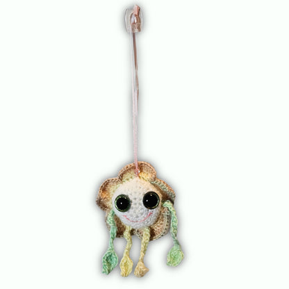 A handmade crocheted frog ornament with large black eyes, a brown and cream body, and green and yellow dangling limbs, hanging from a clear plastic hook against a white background.