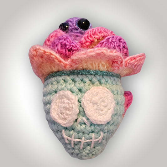 Paisley Peacebones handmade crochet skull flower keepsake with a mint-green skull, white eye sockets, layered pink and purple petals, and a tiny purple sprite peeking from the top.