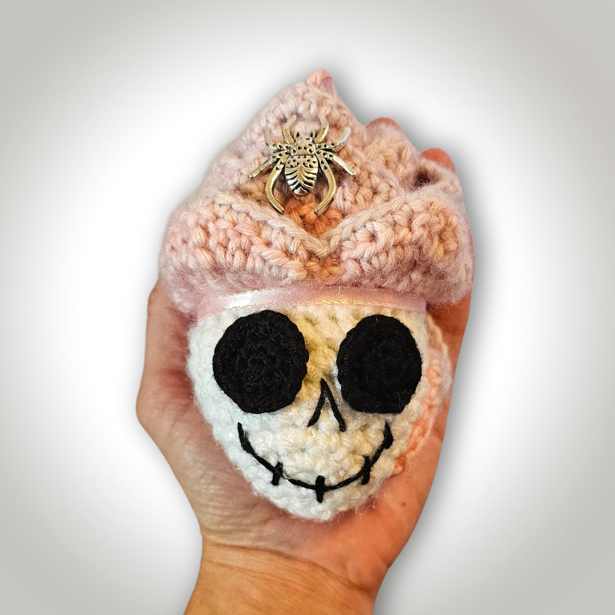 Skelly Rose resting in a hand, showing her white crochet skull face, black stitched features, layered pink flower headpiece, and metallic spider detail.