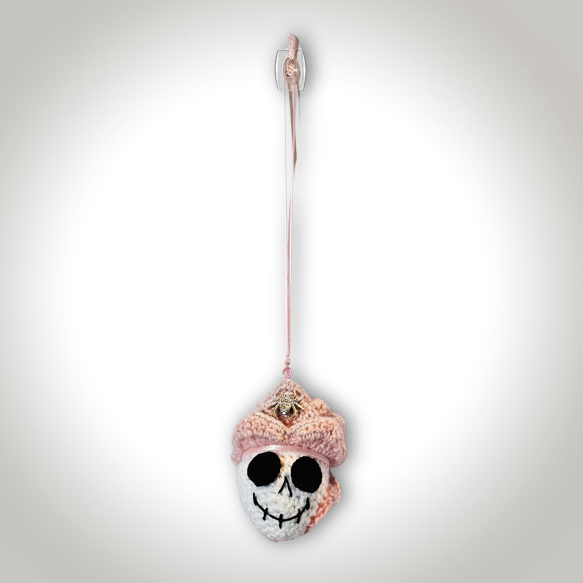 Full front view of Skelly Rose hanging from a pink ribbon as a handmade crochet skull keepsake with layered flower petals and spider detail.