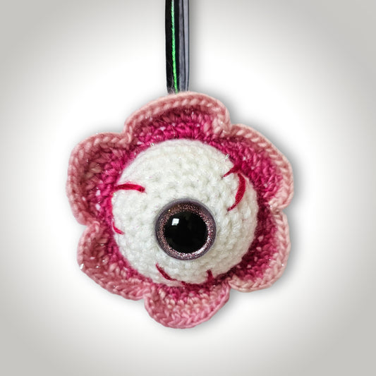 Wanda Winkless handmade crochet eyeball keepsake with a rounded white eye, pink sparkly iris, red stitched veins, and layered pink flower-like edging.
