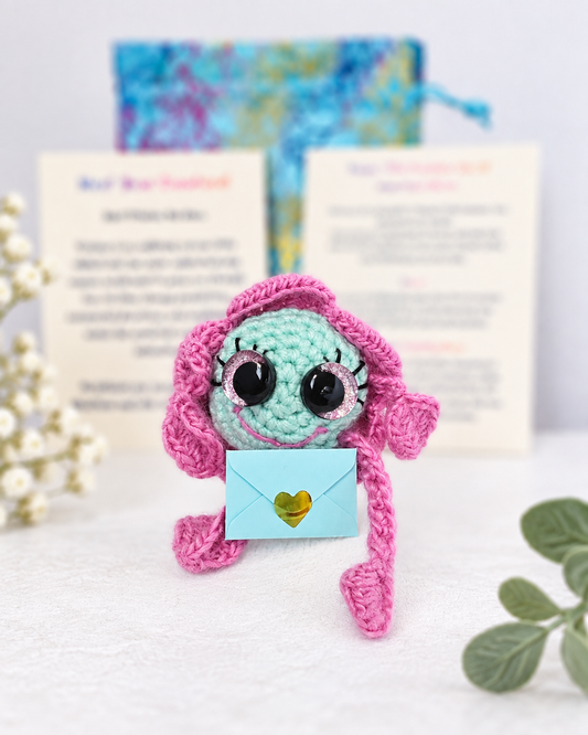 Handmade pink and mint green crochet creature named Lopsy Lucy Pinkmint Juicy holding a small light blue mystery message envelope.