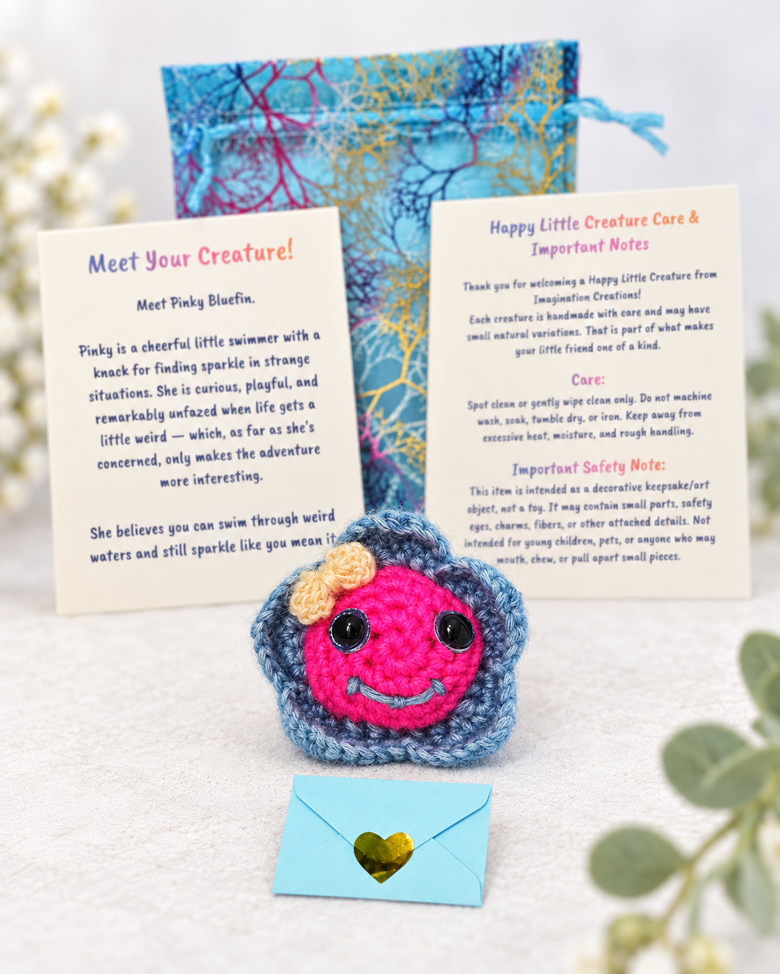 Handmade pink and blue crochet fish creature named Pinky Bluefin with a yellow bow holding a small light blue mystery message envelope.