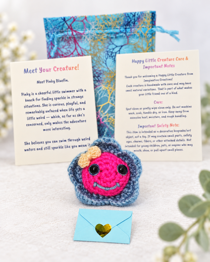 Handmade pink and blue crochet fish creature named Pinky Bluefin with a yellow bow holding a small light blue mystery message envelope.