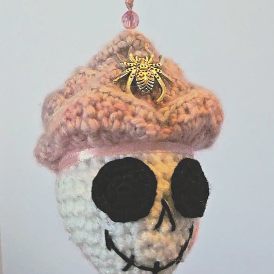 360-degree view of Skelly Rose, a handmade crochet skull keepsake with a sparkly white skull face, layered pink-to-purple flower petals, metallic spiders, bead details, a hidden black sprite, and a peace-sign-inspired back.