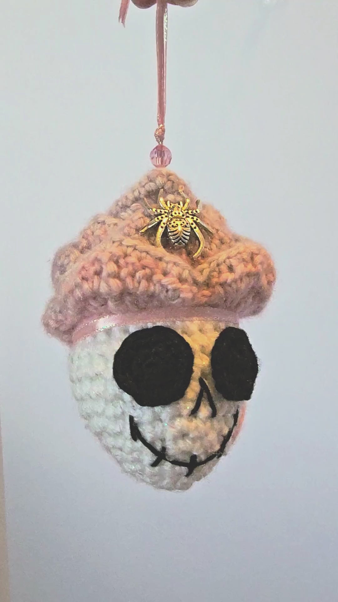 360-degree view of Skelly Rose, a handmade crochet skull keepsake with a sparkly white skull face, layered pink-to-purple flower petals, metallic spiders, bead details, a hidden black sprite, and a peace-sign-inspired back.