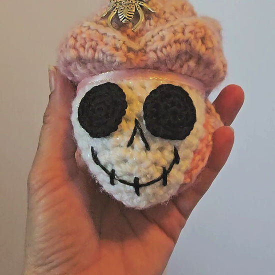 Close-up video of Skelly Rose, a handmade crochet skull keepsake with a sparkly white skull face, layered pink and purple flower petals, metallic spiders, beads, and a hidden black sprite with pink sparkly eyes.