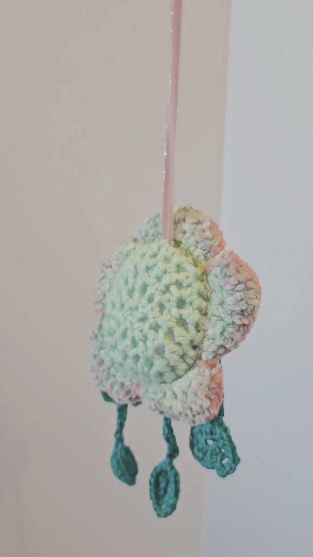 360-degree view of Floyd Flower Boy, a handmade crochet flower keepsake with a mint green face, pastel flower petals, teal dangling limbs, sparkly eyes, and a pink hanging ribbon.