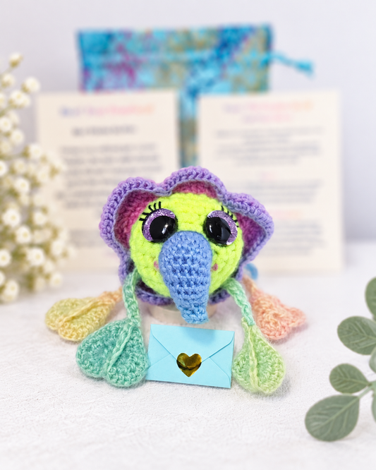 Handmade crochet creature named Tuck Tangletrunk with a long blue trunk and purple hood holding a small light blue mystery message envelope.